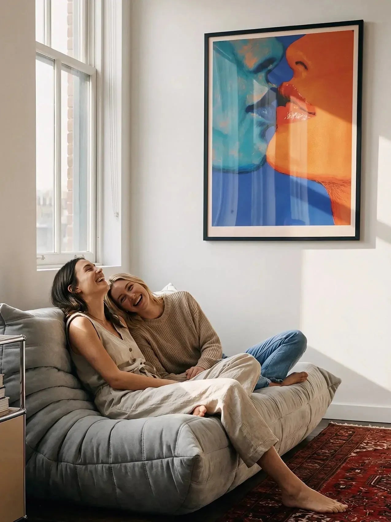Colorful kiss art poster on wall above cozy sofa with two women laughing in bright living room