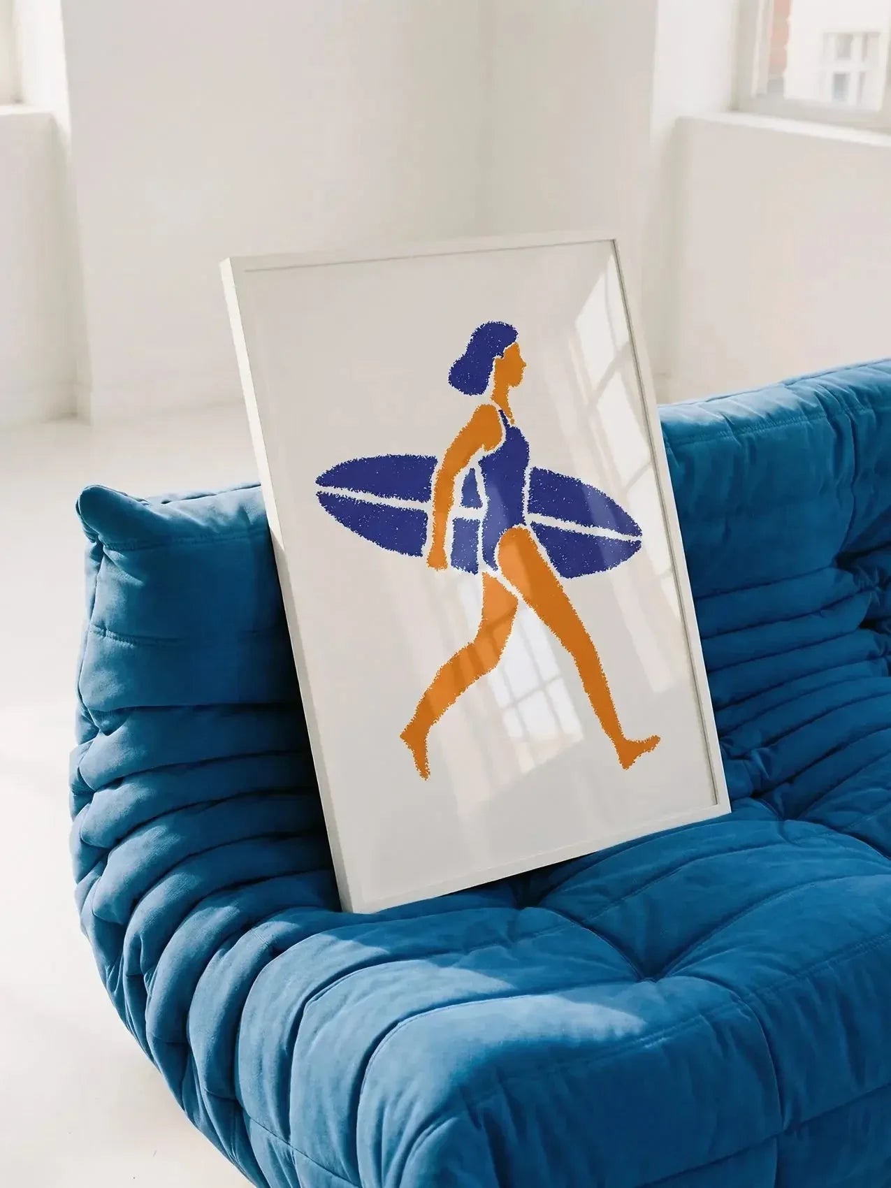Framed surf art print of a woman carrying a surfboard, placed on a blue couch in a bright room.